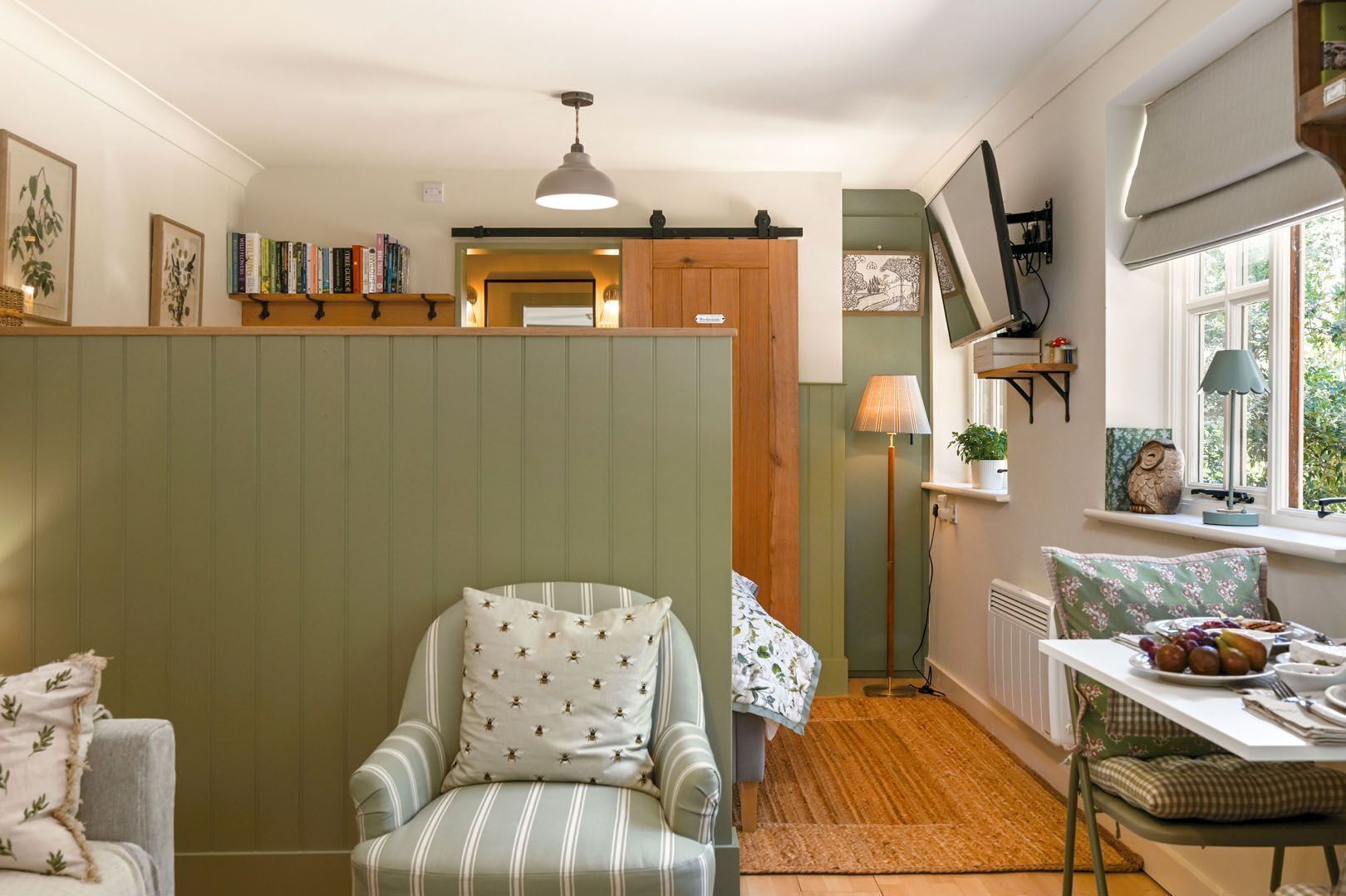A cozy room with a striped armchair, a dining table with a plate of fruit, a lamp, and a bookshelf.
