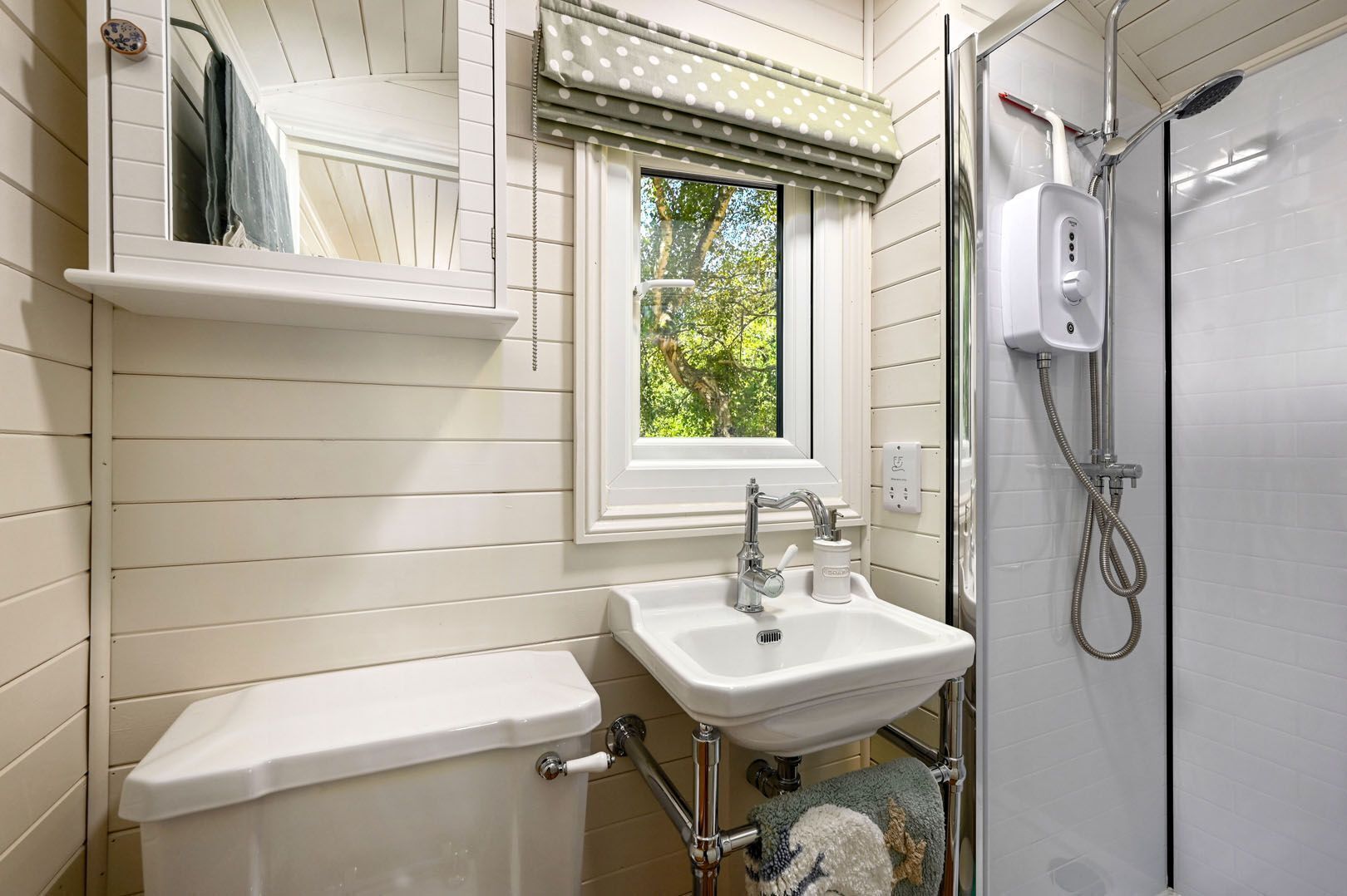 A bathroom with a white sink, a toilet, a window with a polka dot valance, and a shower area.