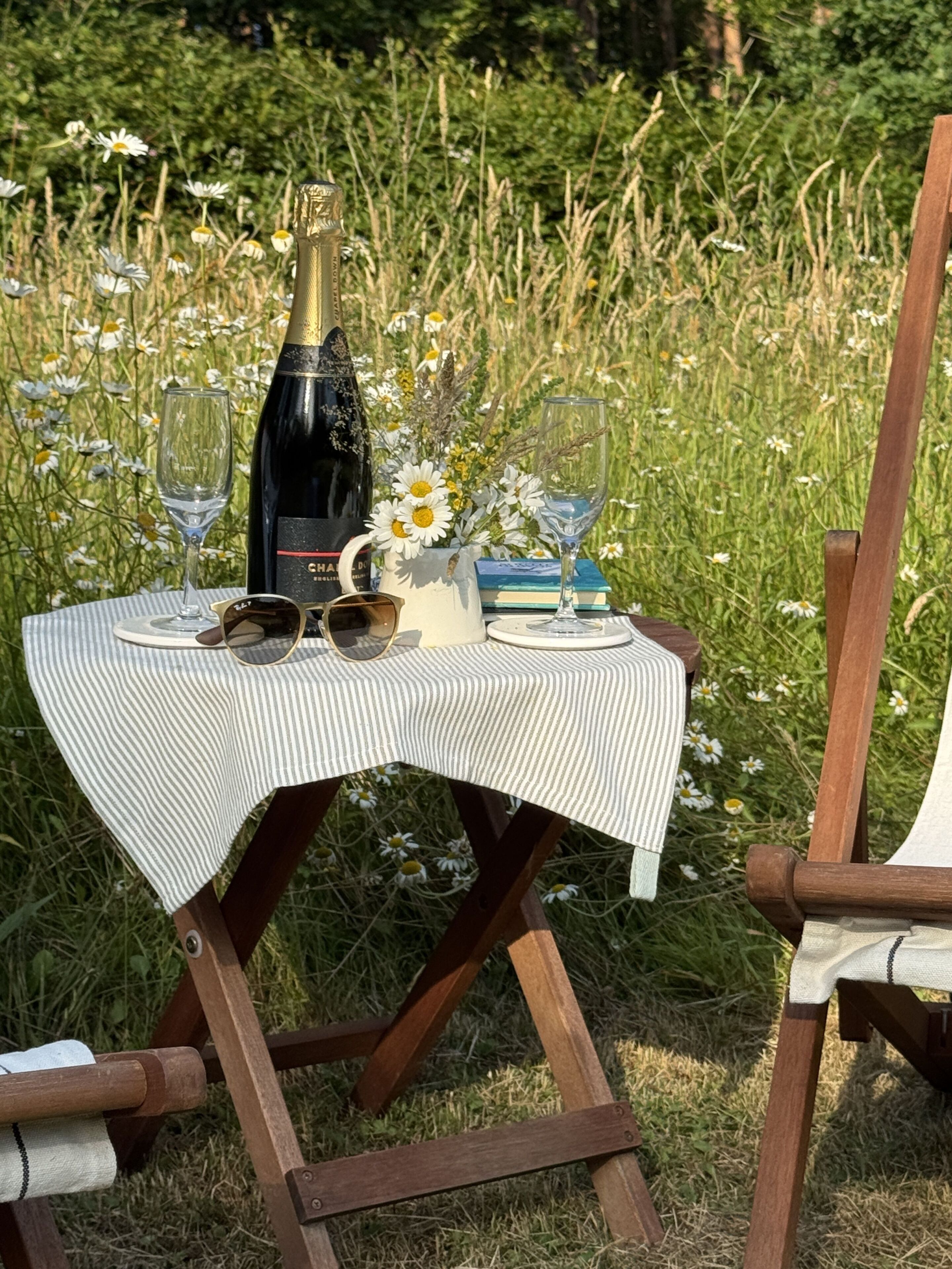 A picnic setup with a bottle, glasses, and a book on a table in a grassy area.