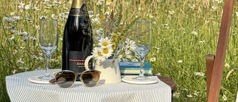 Un àpat preparat per a picnic amb una ampolla, gots i un llibre damunt d'una taula en una zona enjardinada.