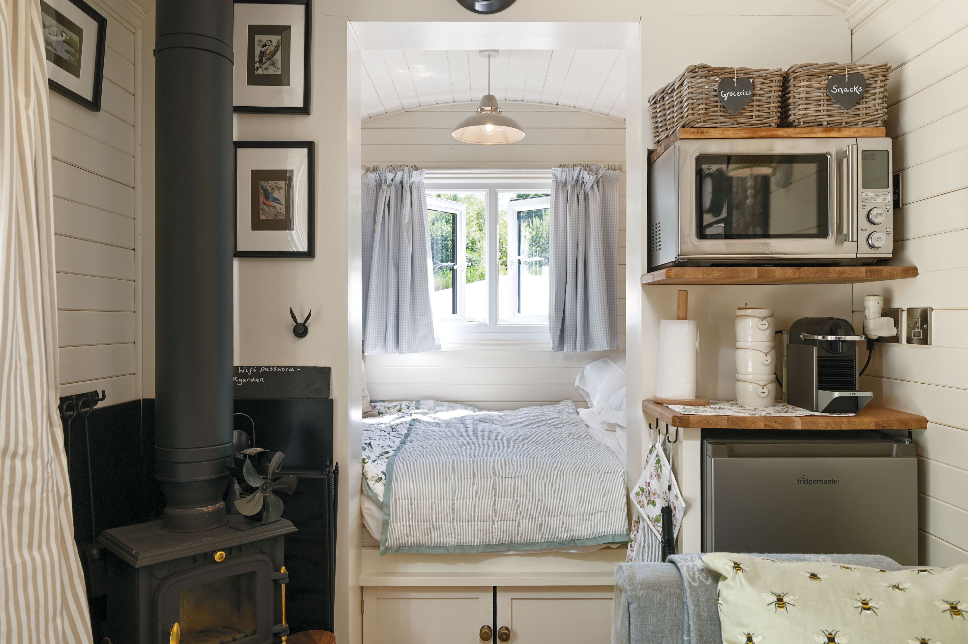A compact bedroom with a bed, a window with curtains, a stove, and a kitchenette with a microwave and kettle.