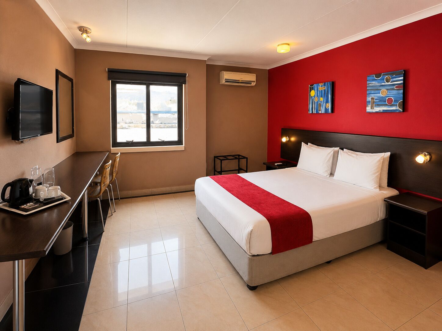 A hotel room with a bed, bedside tables, a desk with a chair, a television, and artwork on the walls.