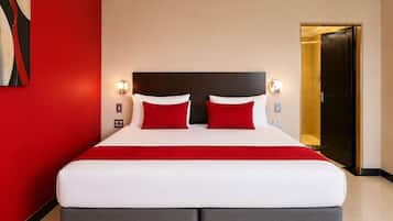 Una habitación de hotel con una cama grande, ropa de cama roja y blanca, un cabecero oscuro y dos lámparas fijadas a la pared.