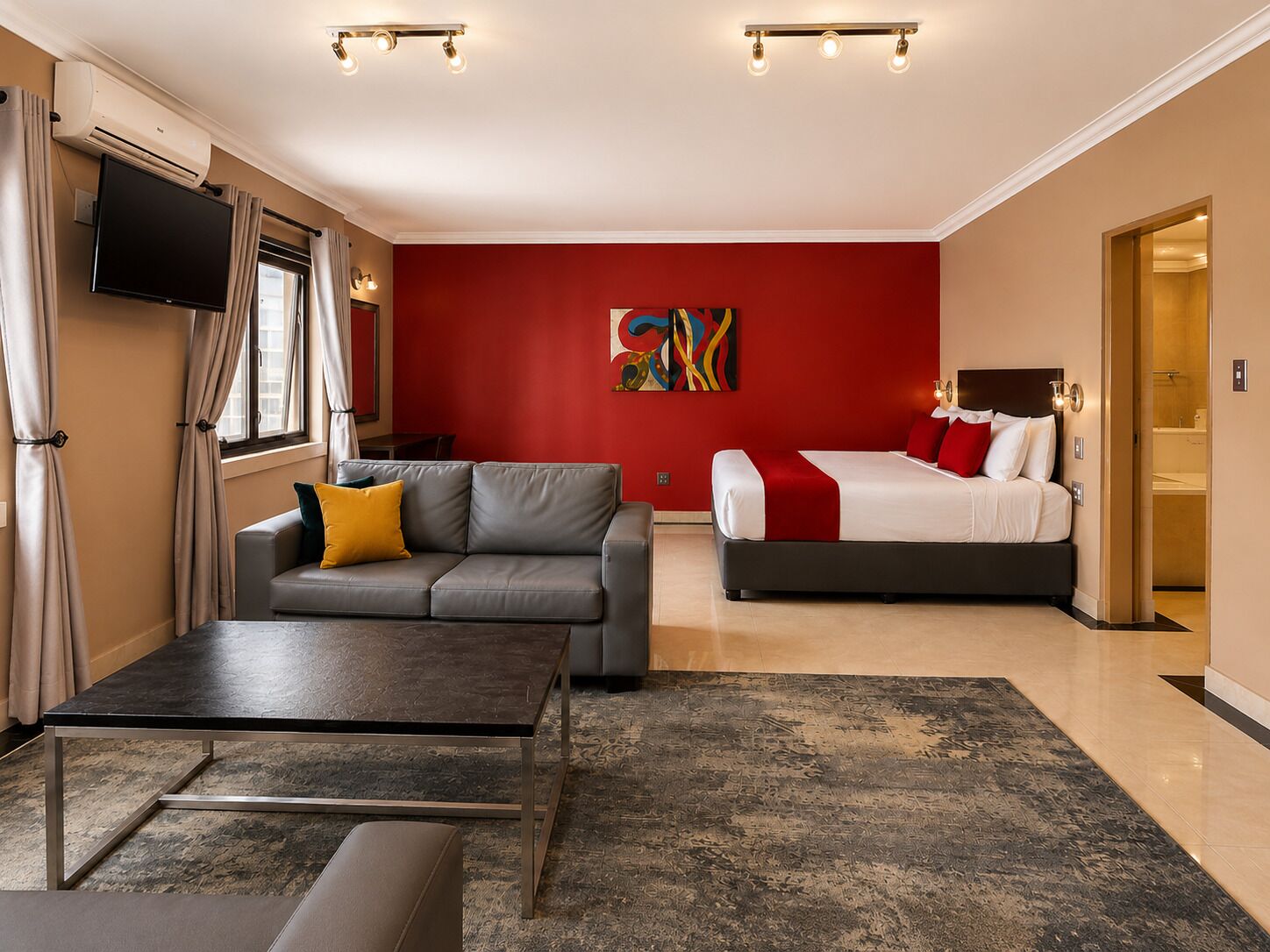Habitación de hotel con un sofá gris, una cama con ropa de cama roja y blanca, un televisor de pantalla plana montado en la pared y un cuadro abstracto grande en la pared roja.