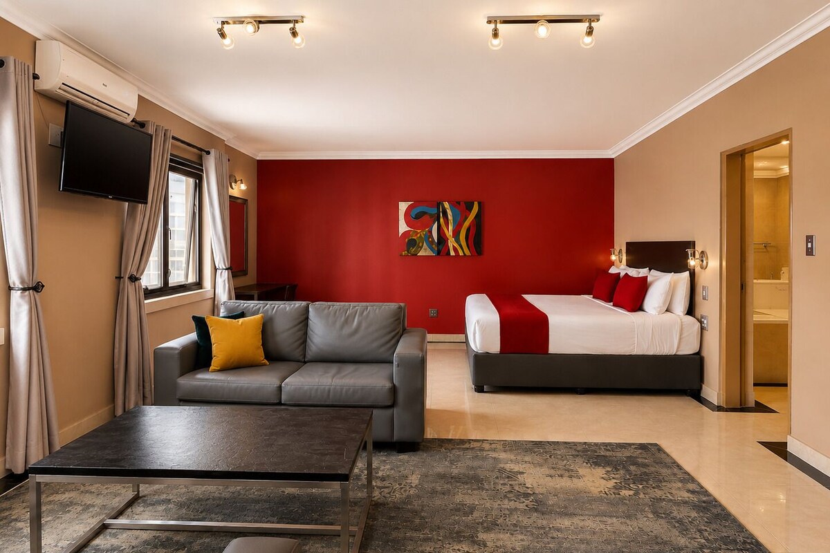 Habitación de hotel con un sofá gris, una cama con ropa de cama roja y blanca, un televisor de pantalla plana montado en la pared y un cuadro abstracto grande en la pared roja.