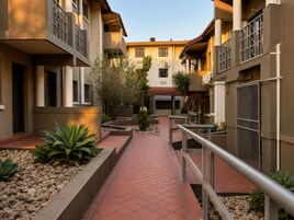 A courtyard with a tiled walkway, planters with various plants, and a building with balconies and railings.
