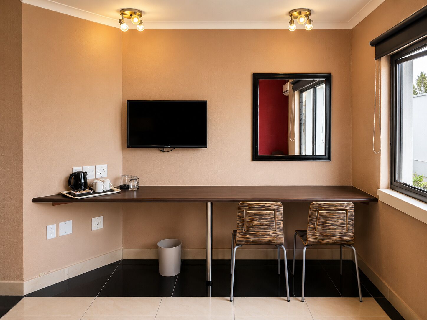 Quarto de hotel com televisão de tela plana na parede, uma mesa pequena com uma cafeteira e uma chávena, duas cadeiras, um cesto de lixo e uma janela com cortina vermelha.