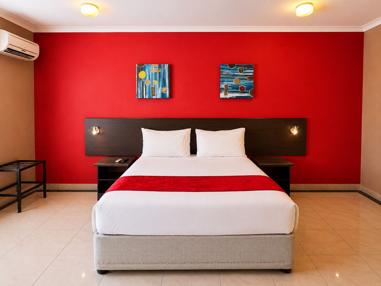 Habitación de hotel con pared roja, una cama grande con ropa blanca, dos mesitas de noche y una obra de arte sobre la cama.