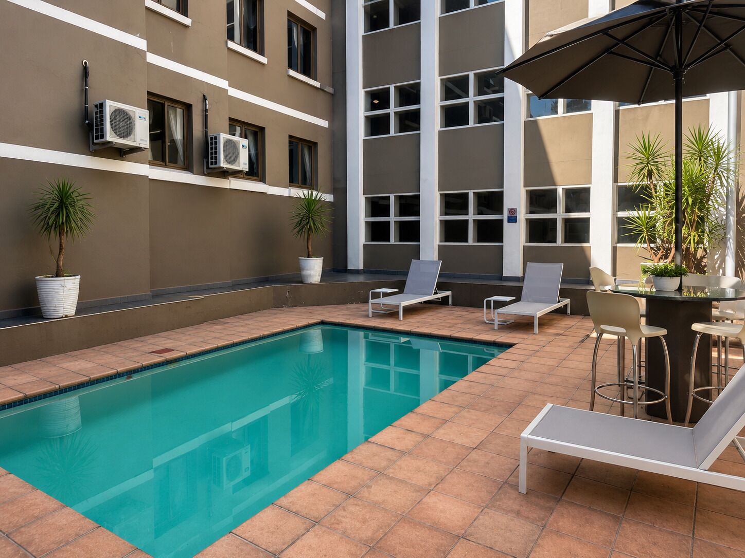 A swimming pool with lounge chairs and a table, surrounded by potted plants and air conditioning units on the building facade.
