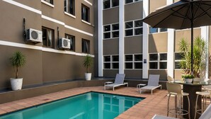A swimming pool with lounge chairs and a table, surrounded by potted plants and air conditioning units on the building facade.