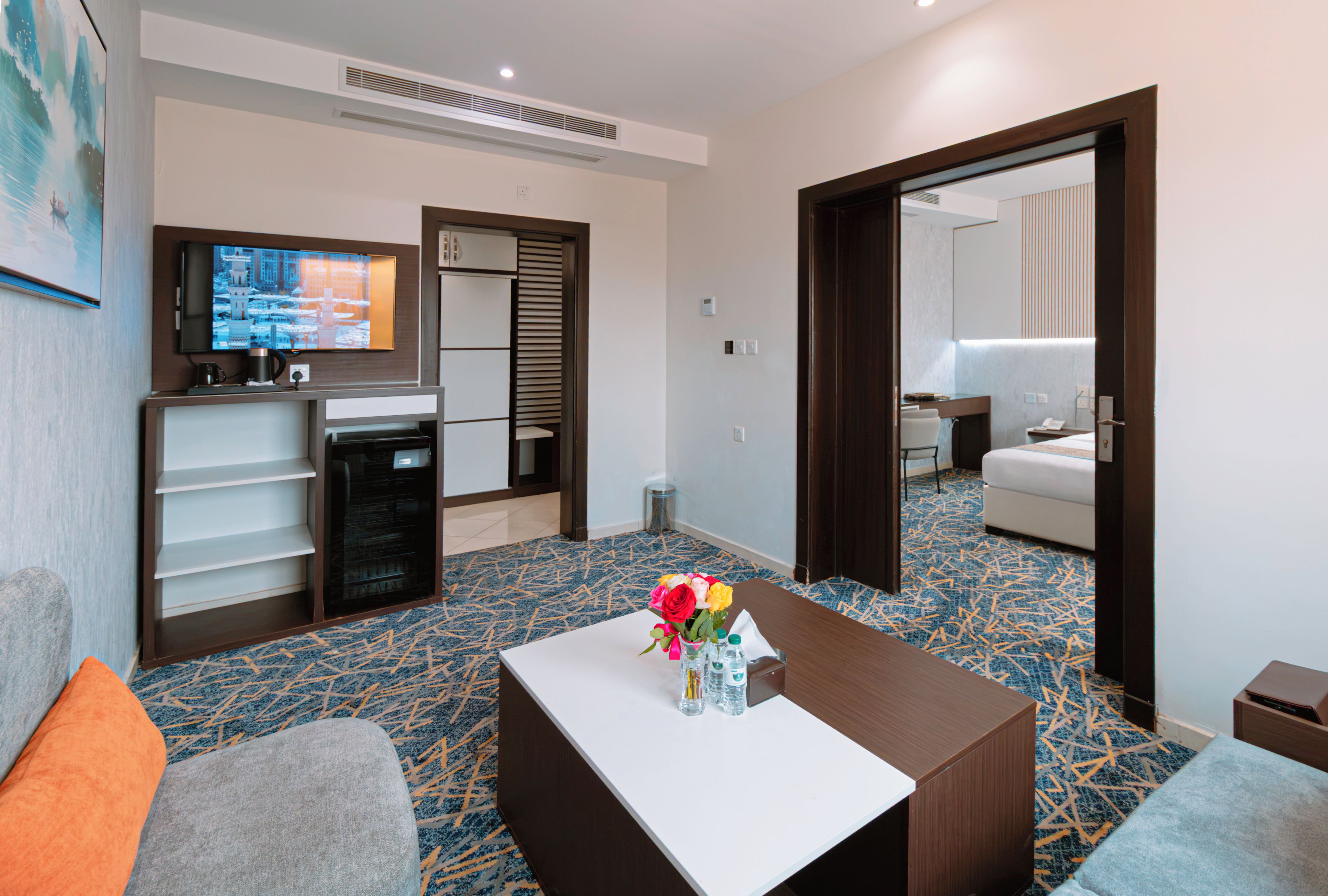 Quarto de hotel com televisão de tela plana na parede, frigobar, escrivaninha com um vaso de flores e carpete estampado.