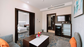 A hotel room with a sofa, a coffee table with a vase of flowers, a television, and a minibar. - Zawaya Almasiya Alawali Apartments (Madinah)
