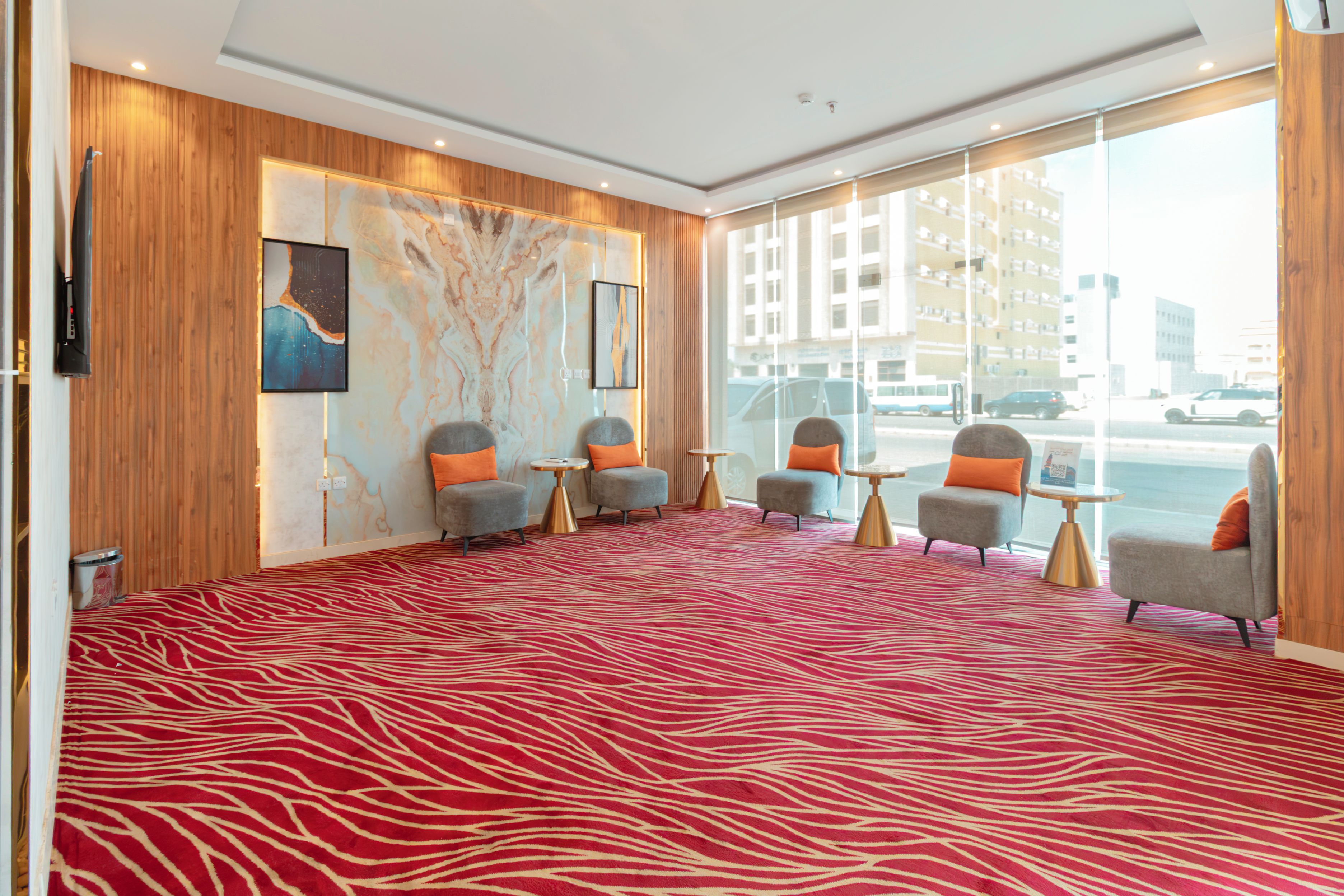 Een hotel lobby met een roodgekleurd tapijt met patroon, vier grijze fauteuils met oranje kussens, een marmeren muur met kunstwerken, en grote ramen met uitzicht op de stad.