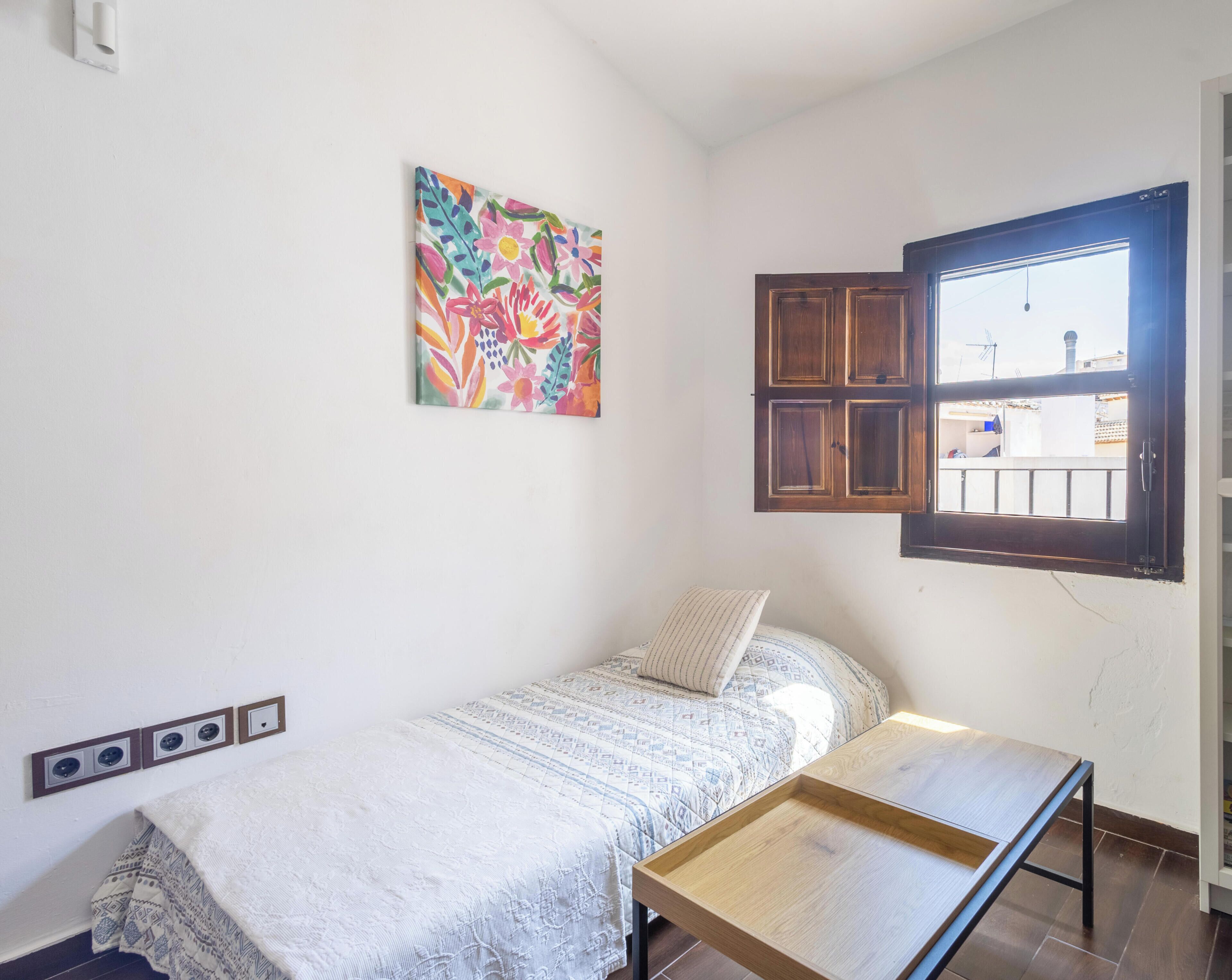 Una habitación pequeña con cama individual, ventana, mesita de noche y un cuadro colorido en la pared.
