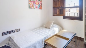 Una habitación pequeña con cama individual, ventana, mesita de noche y un cuadro colorido en la pared.