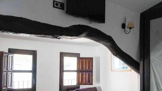 Una habitación con techo inclinado, una cama, una silla con cojines, una mesita y dos ventanas.