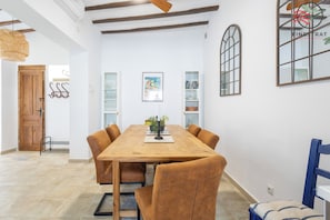 A dining area with a wooden table and brown upholstered chairs, a white wall with a framed painting, and a window with a grid pattern.