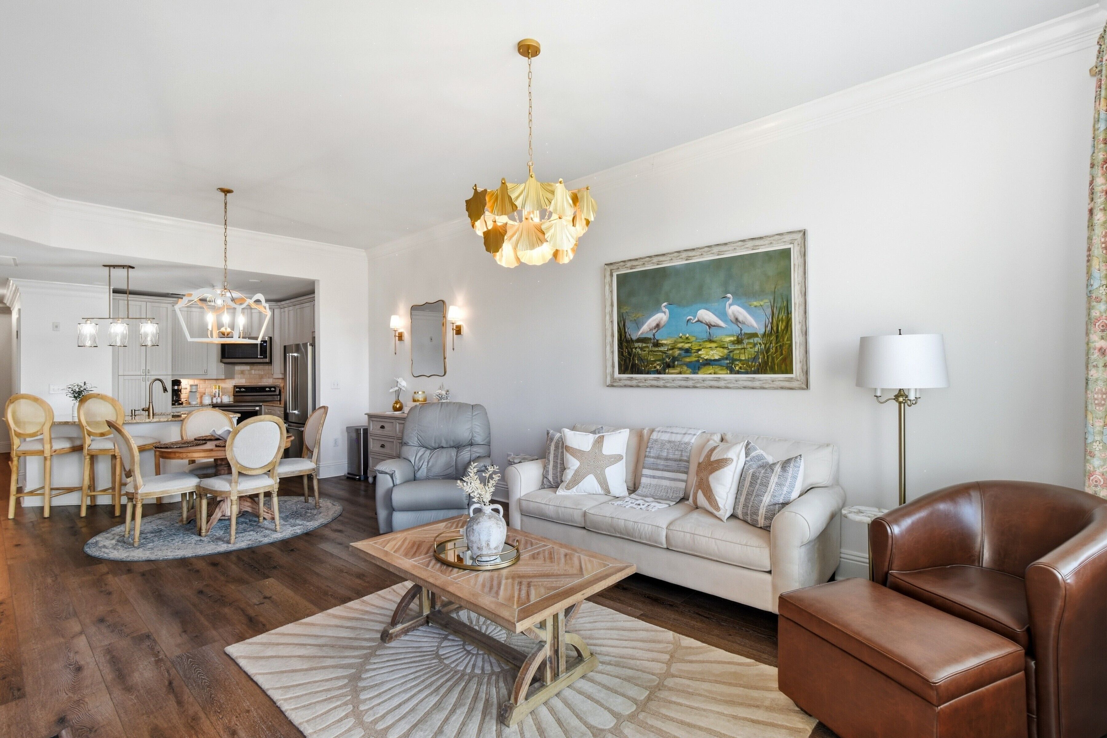 A modern living room with a dining area, a gray sofa, a brown leather chair, a wooden coffee table, and a chandelier.