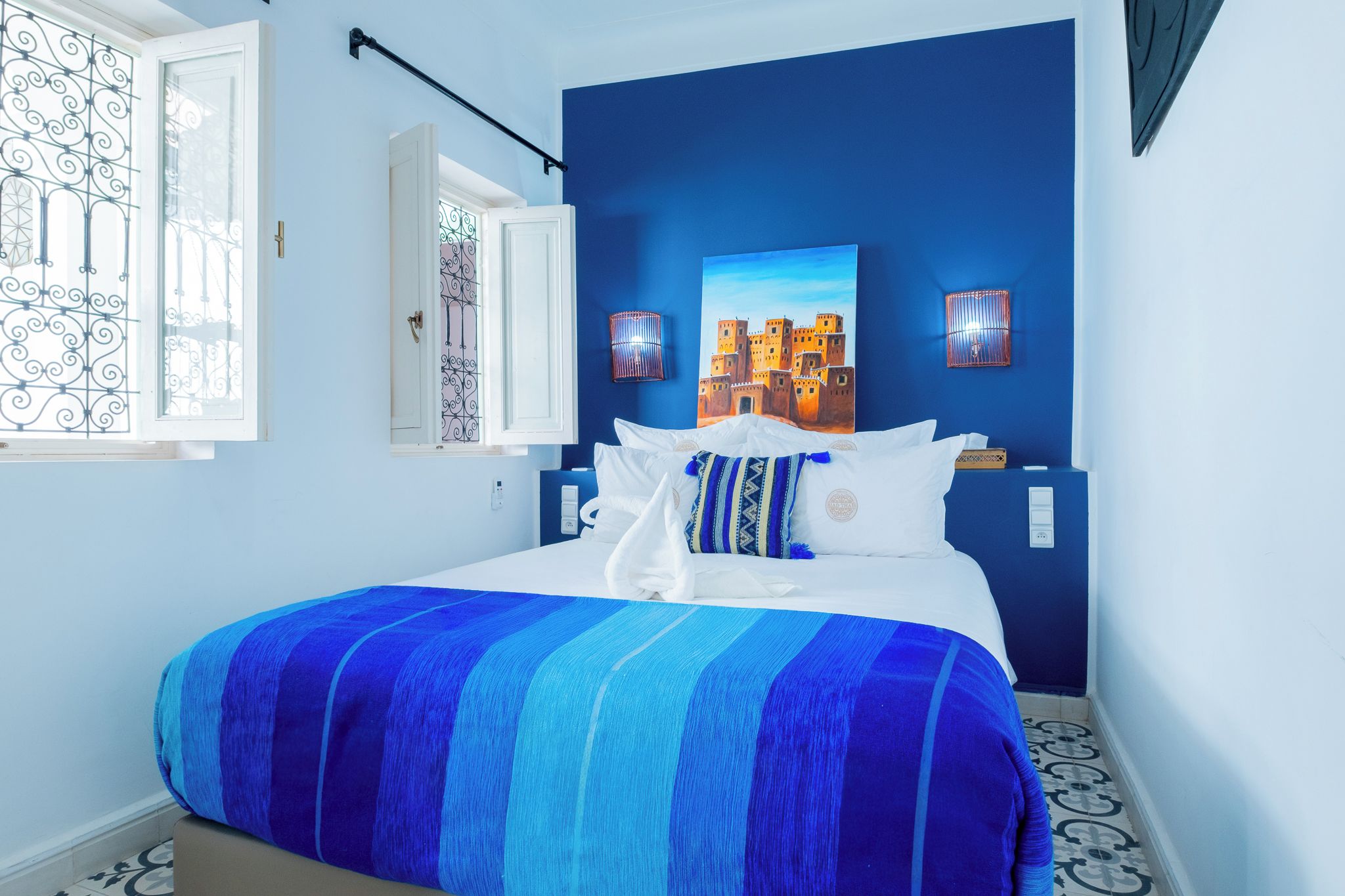 A bedroom with a bed, a window with a decorative grille, a wall-mounted light fixture, and a painting of a castle.