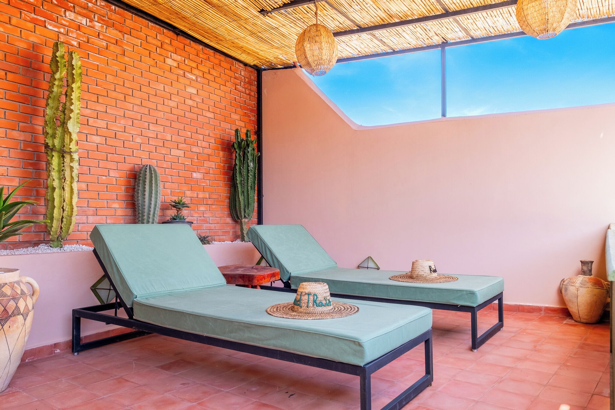 A rooftop lounge area with two green lounge chairs, a woven ceiling, and cacti against a brick wall.