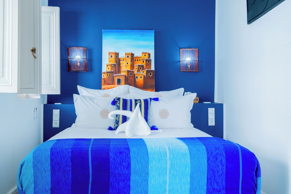 A hotel room with a bed, a painting of a castle, two wall sconces, and a door.