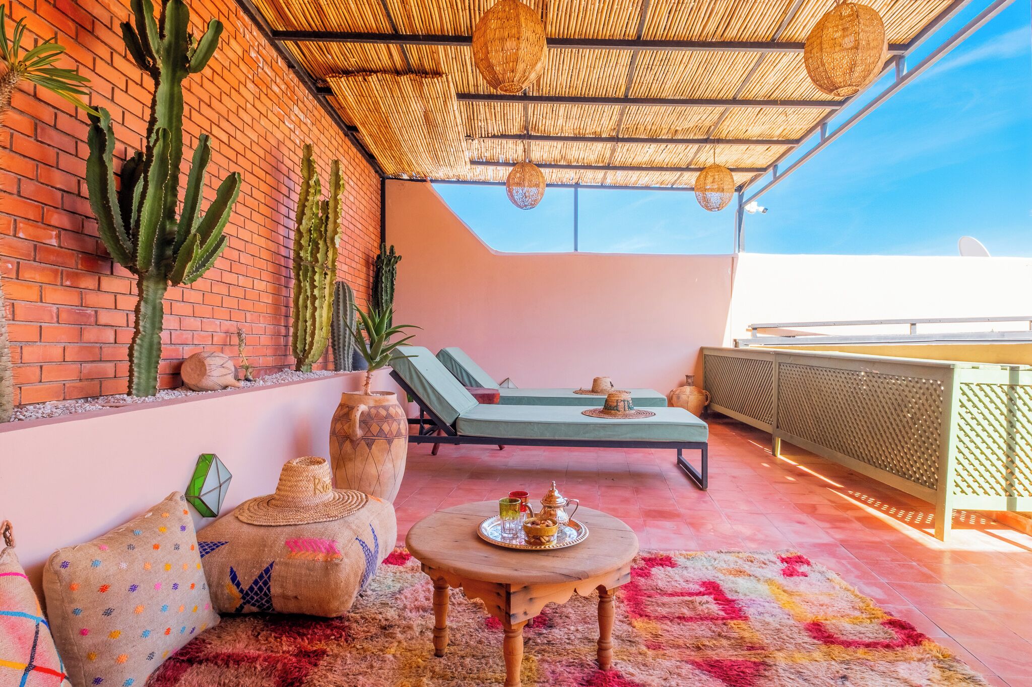 A rooftop terrace with a brick wall, cacti, lounge chairs, a round wooden table with a tray, and a woven ceiling.