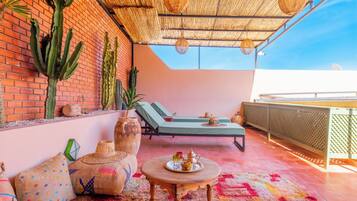 A rooftop terrace with a brick wall, cacti, lounge chairs, a round wooden table with a tray, and a woven ceiling.