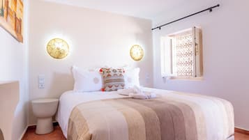 Quarto com cama, mesas de cabeceira, janela com grades de ferro decorativas e luminárias fixadas na parede.