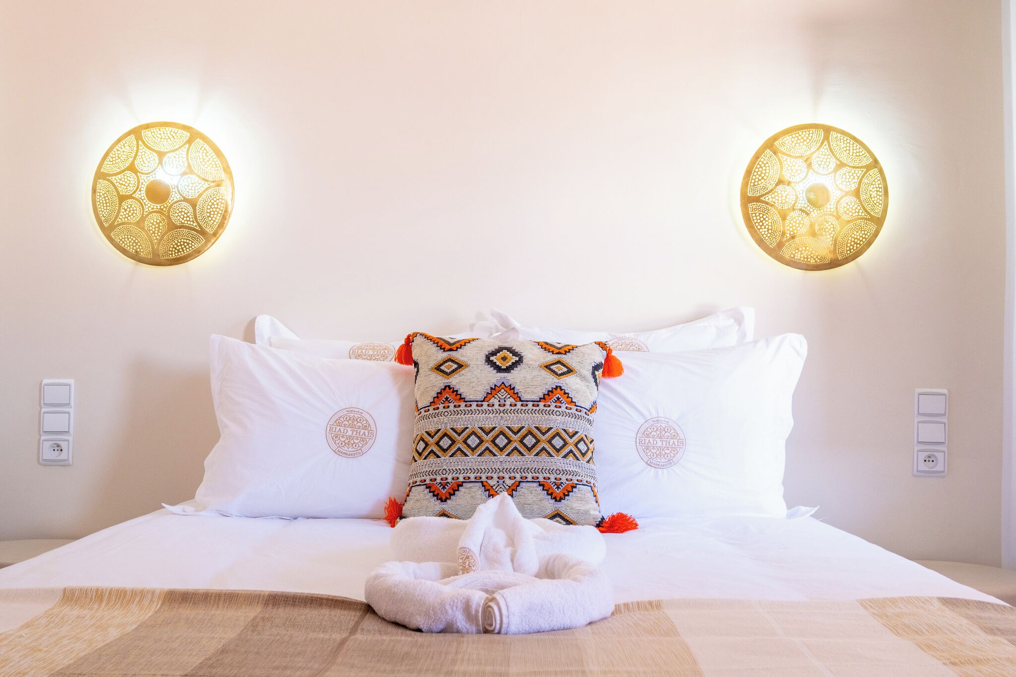 A hotel room with a bed, two decorative wall sconces, and a patterned throw pillow.