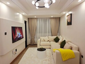 Una sala de estar moderna con un sofá color beige, un televisor de pantalla plana, una mesa de centro redonda y un ventanal con cortinas.