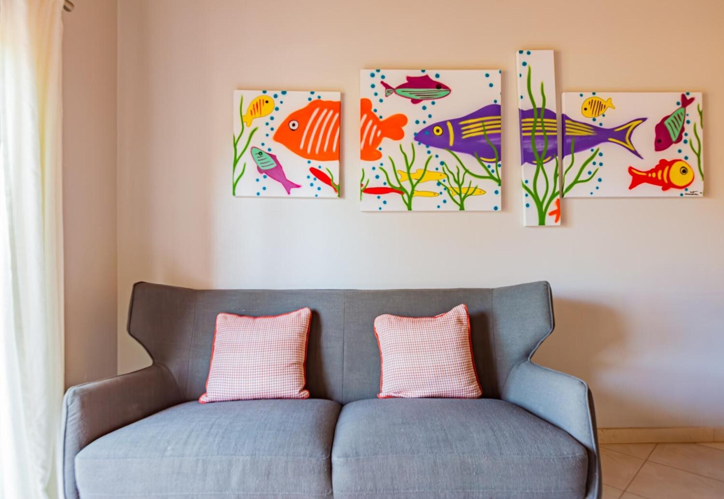 A grey sofa with two checkered cushions against a wall with three colorful fish paintings.
