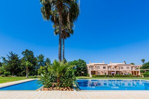 A swimming pool with a palm tree in the foreground, a large building with a unique architectural style in the background, and a clear blue sky. - 4-Bedroom Villa near the Beach – Casa Colina 2 (Albufeira)