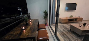 Ein Essbereich mit einem Holztisch, zwei Stühlen und Kerzen. Eine Topfpflanze und ein an der Wand befestigter Fernseher sind ebenfalls vorhanden.