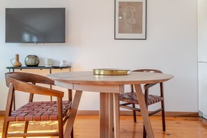 Un coin repas avec une table ronde en bois, deux chaises à assise en osier, un téléviseur à écran plat fixé au mur et une œuvre d’art encadrée au-dessus de la table.