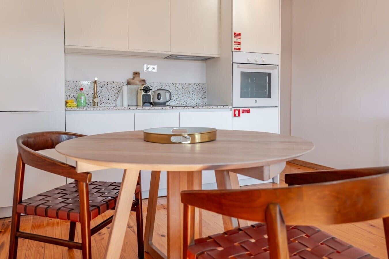 Una cocina compacta con gabinetes blancos, horno integrado y una mesa redonda de madera con sillas.