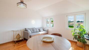 Amplia habitación con una mesa redonda de madera, un sofá gris, una mesita auxiliar con un florero y un gran ventanal con vistas al campo.