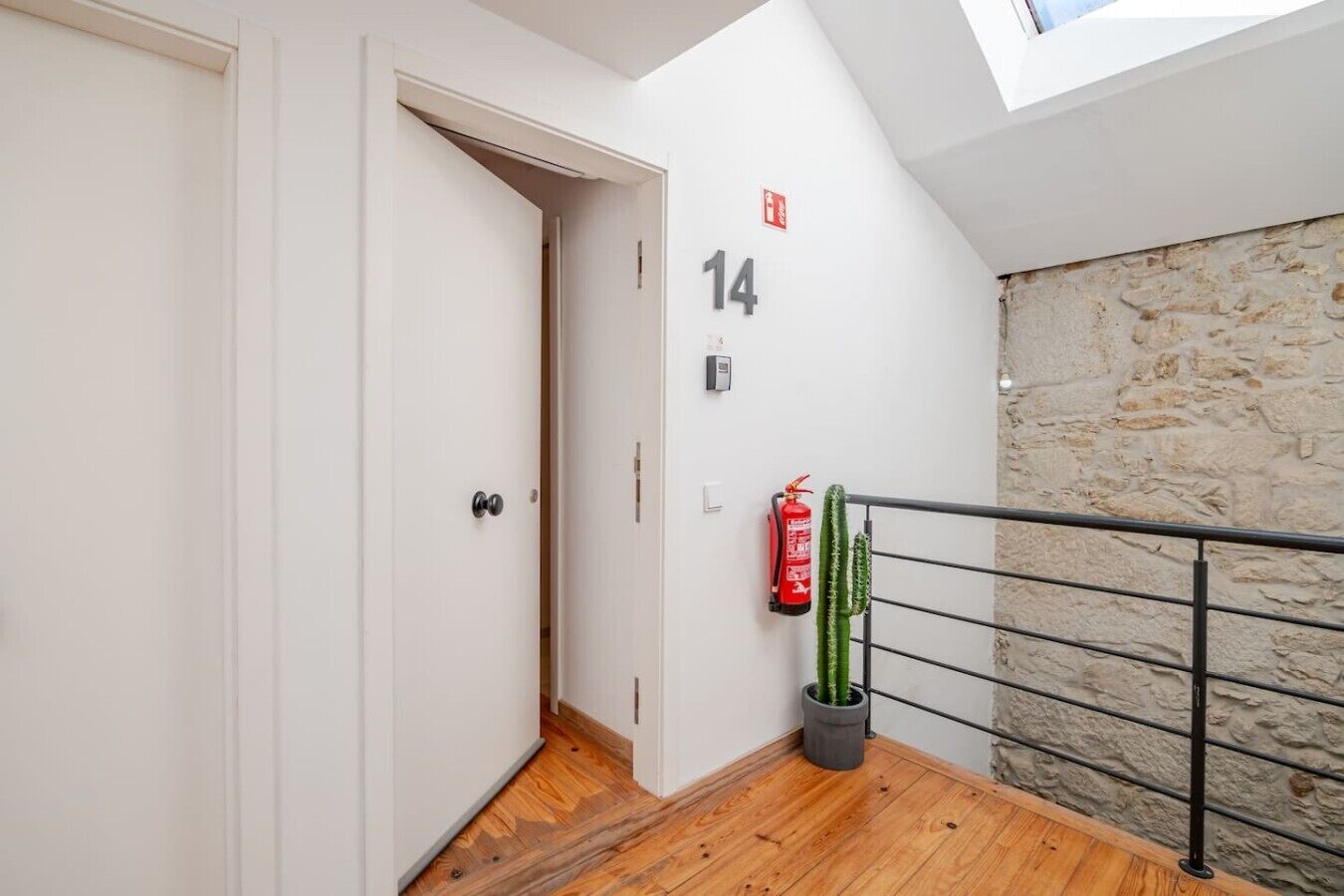 Un couloir avec un plancher en bois, une porte blanche, une plaque portant le numéro « 14 », un extincteur et un cactus dans un pot.