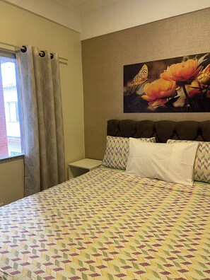 Habitación de hotel con cama, mesita de noche, ventana con cortinas y un cuadro de flores colgado en la pared.