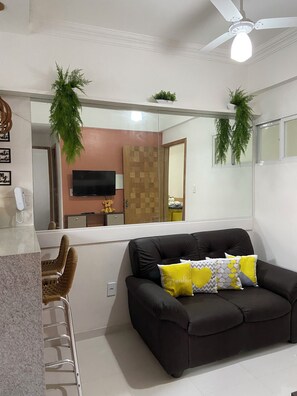 Sala de estar com um sofá preto, uma mesa de centro de vidro e uma televisão. Há um armário de madeira e um espelho na parede.