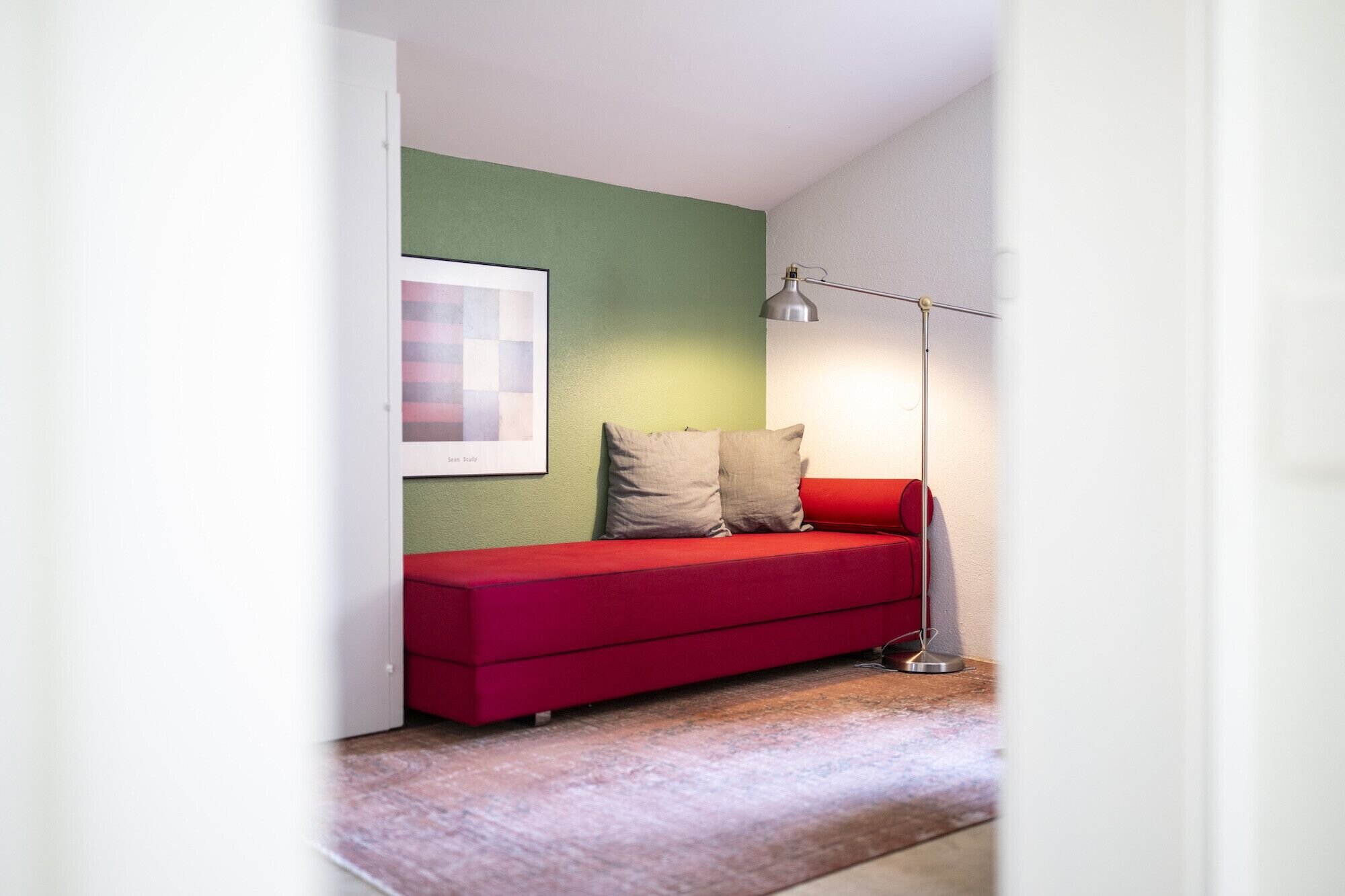 Ein rotes Sofa mit zwei Kissen vor einer grünen Wand, eine Stehlampe und ein gerahmtes Bild.