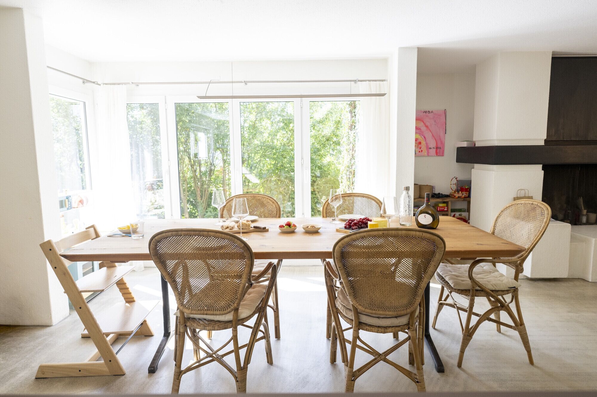 Un espace repas avec une longue table, des chaises en osier et une vue sur la verdure grâce à de grandes fenêtres.
