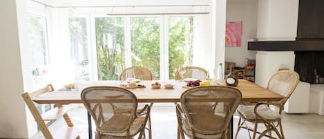 Un espace repas avec une longue table, des chaises en osier et une vue sur la verdure grâce à de grandes fenêtres.