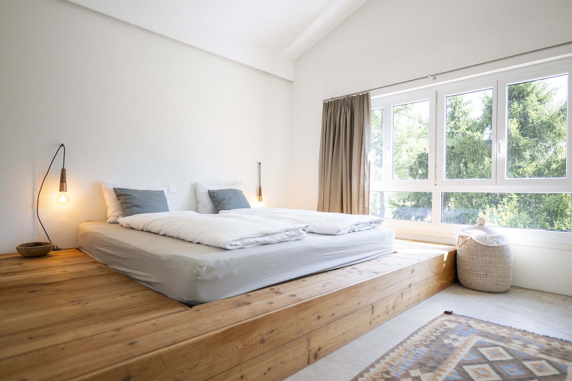 Un dormitorio con piso de madera, una cama con ropa de cama blanca, una lámpara de noche, una ventana con cortinas y vistas a la vegetación exterior.