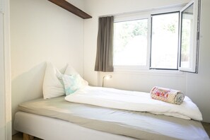 Ein Einzelbett mit Kissen und einer zusammengerollten Decke, ein Fenster mit Vorhängen und ein Holzbalken über dem Bett.