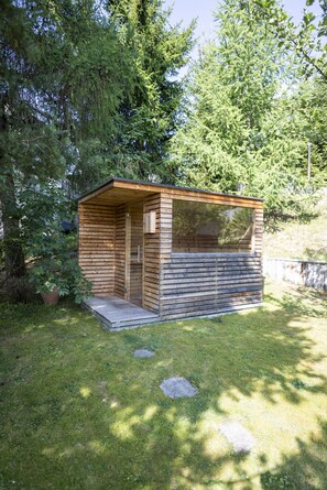 A small wooden cabin with a glass door, surrounded by lush greenery. - Chasa Val 739 - Spacious vacation home with a garden sauna and hot tub in Scuol (Scuol)