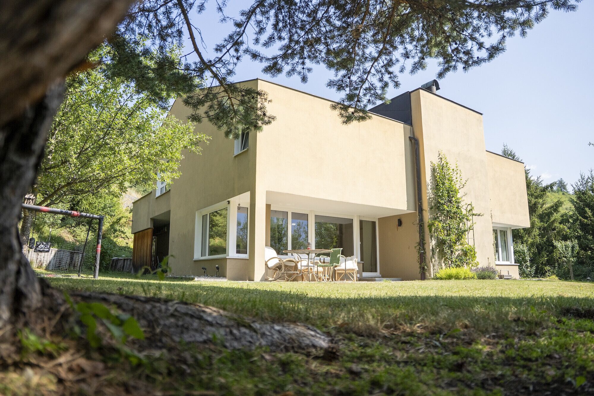 A modern two-story building with a flat roof, large windows, and a patio area with outdoor furniture.