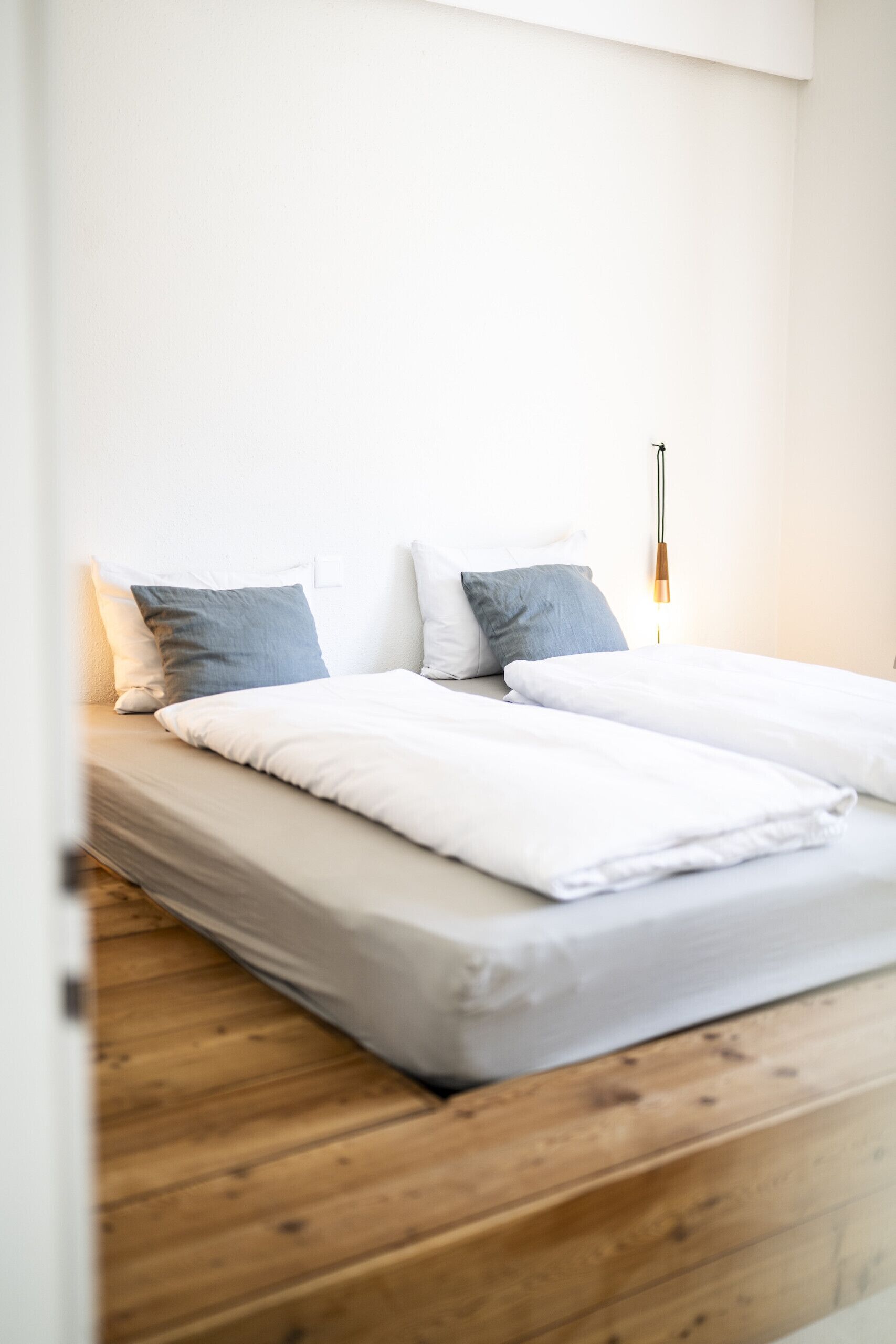 A neatly made bed with white bedding and two pillows, a wooden floor, and a wall-mounted light fixture.