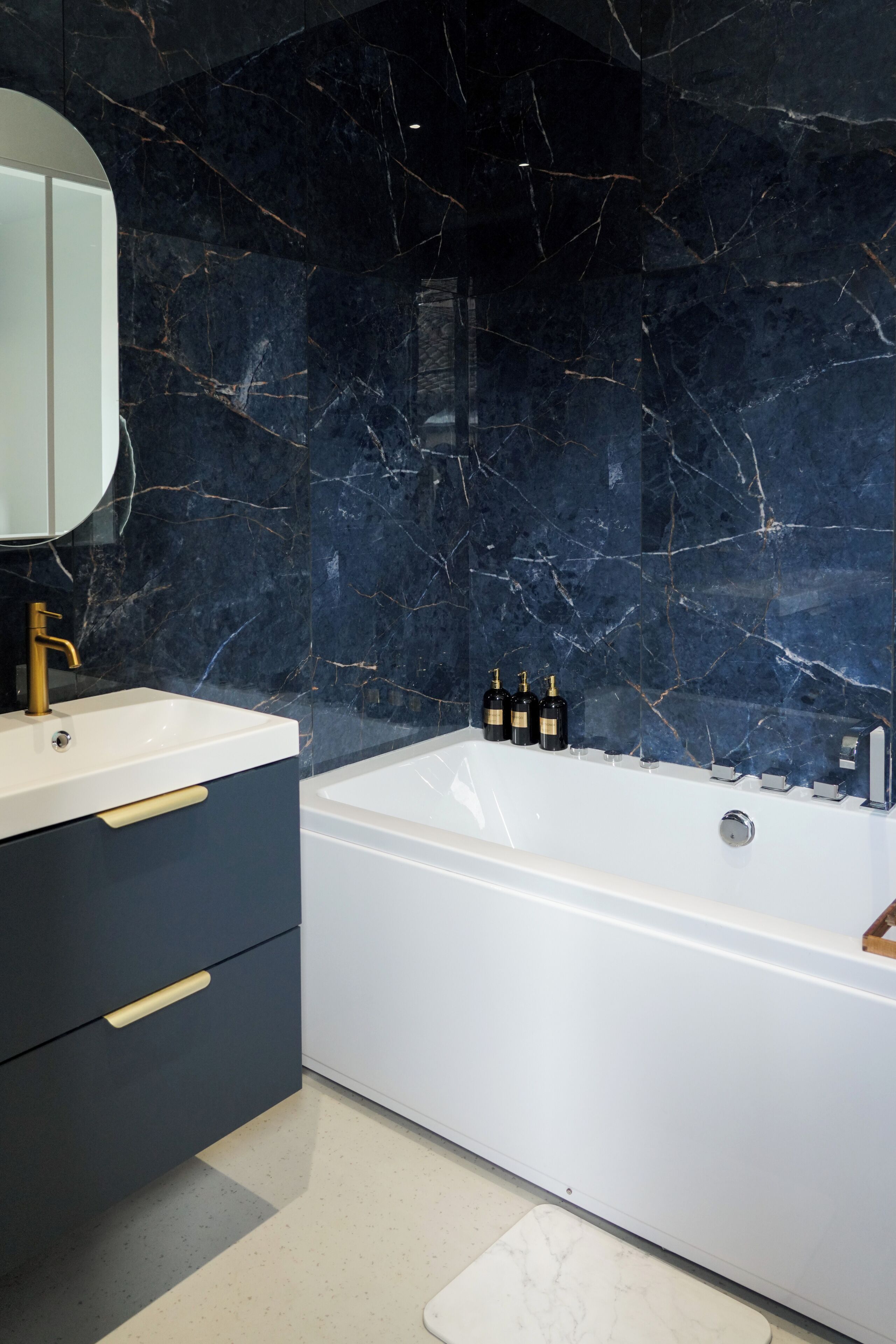 A modern bathroom with a marble wall, a white bathtub, and a vanity with a gold faucet.