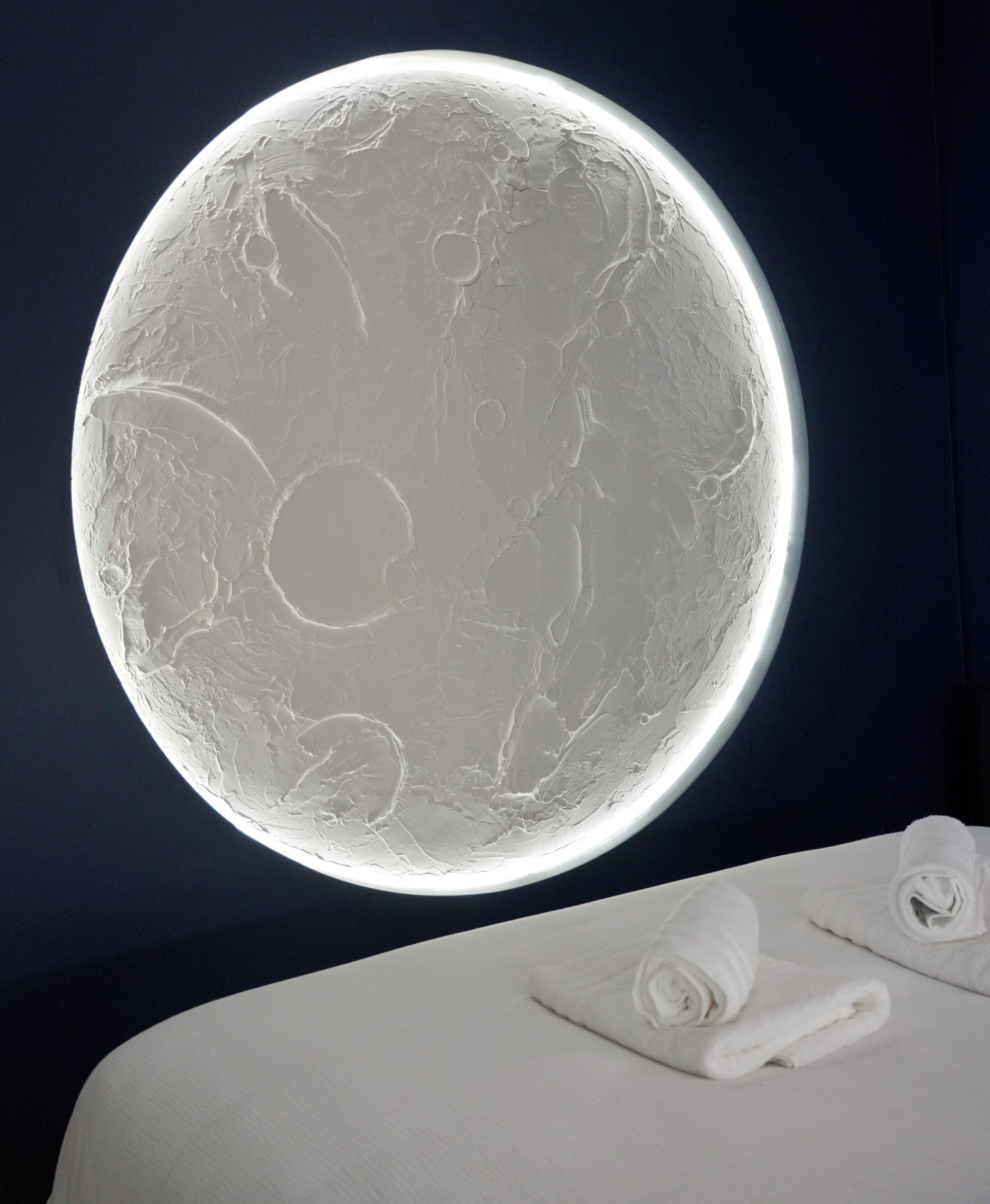 A round, illuminated wall with a textured surface, a bed with white linens, and a dark blue background.