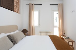 A hotel room with a bed, a bedside table, a lamp, a radiator, and curtains. - Beautiful one-bedroom apartment on Calle Castillo, Madrid, perfect for extended stays. Mediterranean style blends seamlessly with rustic touches of exposed brick and natural wood, creating a warm, bright, and inviting atmosphere. (MADRID)